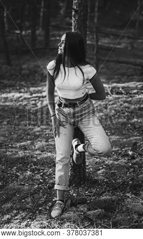 Young Trendy Pretty Woman In Moms Jeans And A White Top Is Leaning And Holding Her Hand On A Tree In