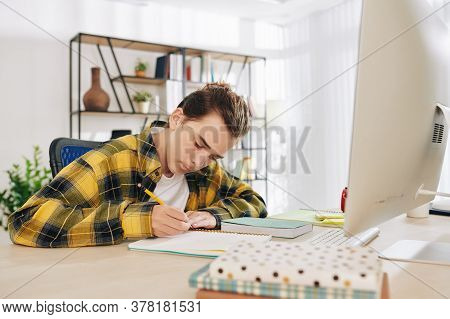 Serious Concentrated Teenger Drawing Or Writing In Notebook When Sitting At His Desk At Home