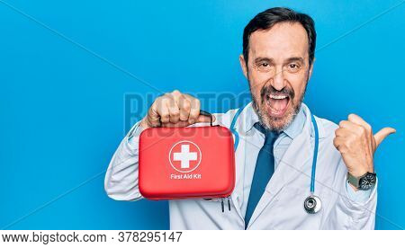 Middle age handsome doctor man wearing coat and stethoscope holding first aid kit pointing thumb up to the side smiling happy with open mouth
