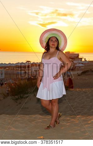 Woman In A White Dress And A Straw Hat On The Beach Against The Background Of The Sea At Sunset.