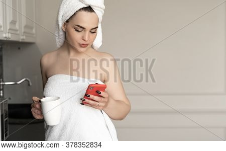 Attractive Caucasian Young Woman With Towel On Head Wrapped In Bathtowel Drinking Tea And Using Smar