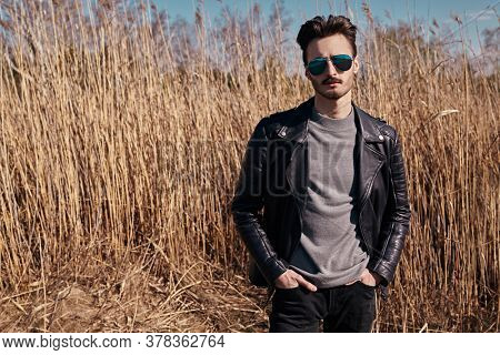 Handsome young man in a black leather jacket and sunglasses stands in the park next to the reeds. The spirit of freedom and adventure. Men's fashion. 