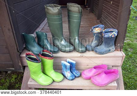 Old Dirty Rubber Boots Of A Big Family Stand On The Wooden Floor Of A Country House Porch Lit By Sum