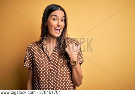 Young beautiful brunette woman wearing casual shirt over isolated yellow background smiling with happy face looking and pointing to the side with thumb up.