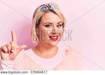 Beautiful blonde plus size woman wearing pincess tiara with diamonds over pink background smiling with an idea or question pointing finger up with happy face, number one