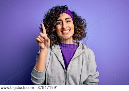 Beautiful curly arab sportswoman doing sport wearing sportswear over purple background showing and pointing up with finger number one while smiling confident and happy.