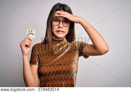 Young beautiful girl holding reminder with sad emotion emoji face over white background stressed with hand on head, shocked with shame and surprise face, angry and frustrated. Fear and upset.