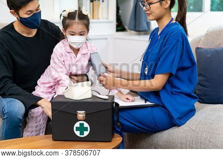 Asian doctor take blood pressure measurement to little girl child sit with her father inliving room while doctor visit home. Home healthcare delivery concept.