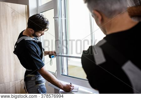Two Professional Workers In Uniform Using Tape Measure While Measuring Window And Making Notes For I