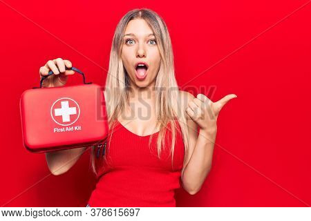 Young blonde woman holding first aid kit pointing thumb up to the side smiling happy with open mouth 