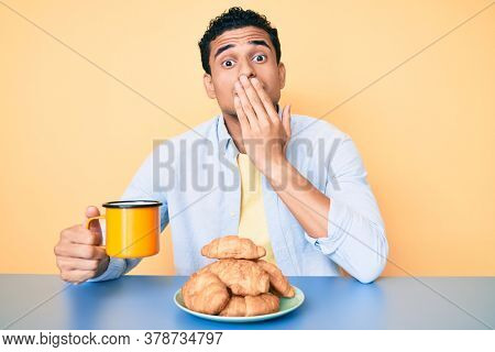 Young handsome hispanic man sitting on the table having breakfast covering mouth with hand, shocked and afraid for mistake. surprised expression 