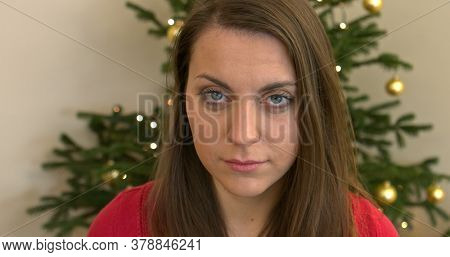 Young calm woman is looking at camera and drinking hot drink, tea or coffee, from mug. She is smiling. Behind her is christmas tree. It's Christmas time.