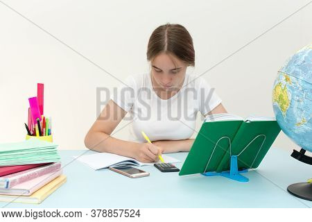 A Girl Sits At A Table And Writes Homework In A Notebook. Overwrites From A Book. Student.student. O