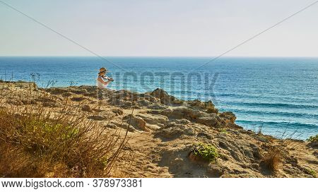 Arsuf, Israel - July 22, 2020: A Girl Sitting On A Hill Above The Sea And Playing The Violin At Suns