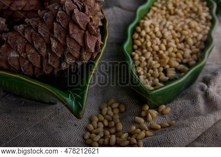 Peeled Pine Nuts And Brown Pine Cone In Green Plates On Old Burlap. Tasty And Healthy Nuts Rich In P