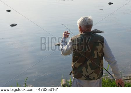 Old Man Fishing. Senior Gray Haired Fisherman Throws A Spinning From Shoreside At Sunset, Twisting A