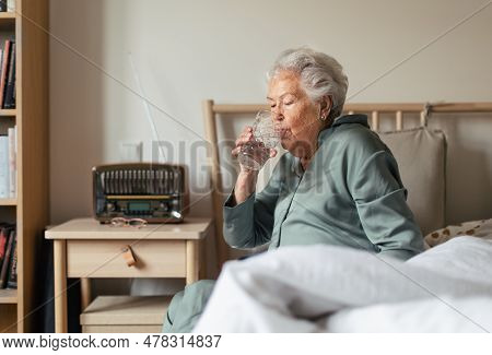 Senior Woman Sitting In Pajama On Her Bed And Drinkig Water.