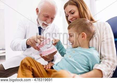 Senior Male Dentist Educating Child Patient Sitting In Mother's Lap About Proper Teeth Brushing Tech