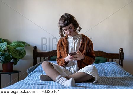 Pensive Dreamy Student Girl Sitting Crosslegged On Bed Looking To Smart Phone Screen, Reading Online
