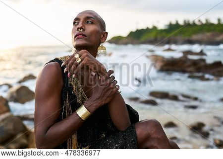 Non-binary Black Person In Luxury Dress Sits On Rocks In Ocean. Trans Ethnic Fashion Model Wearing J