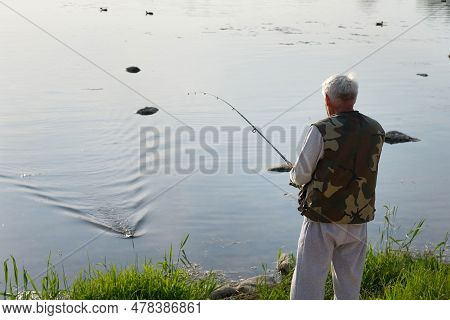Old Man Fishing. Senior Gray Haired Fisherman Throws A Spinning From Shoreside At Sunset, Twisting A