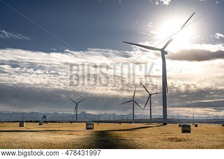 Windmills Sillhouette Standing Tall Overlooking A Harvested Field With Round Bales Of Hay On The Can