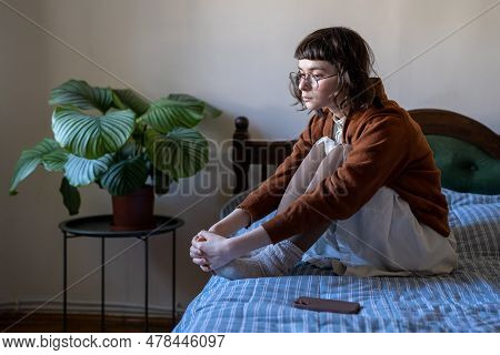 Pensive Sad Young Woman Sits On Bed, Hugs Knees Next To Mobile Phone Waiting For Call, Message, Sms.