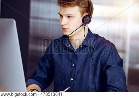 Young Blond Businessman Using Headset And Computer In A Darkened Office, Glare Of Light On The Backg