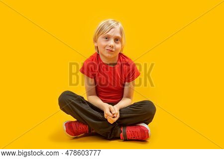 Blond Boy Wears Red T-shirt And Sneakers Sit On The Floor With Crossed Legs Isolation On Yellow Back