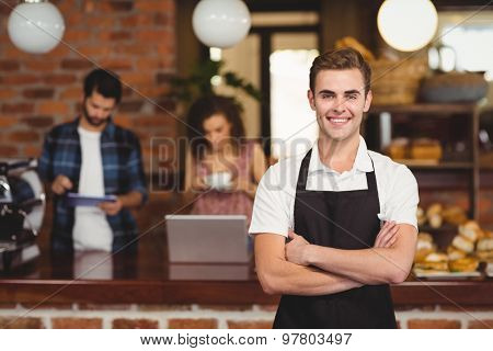 Portrait of smiling barista with arms crossed in front of customers at coffee shop