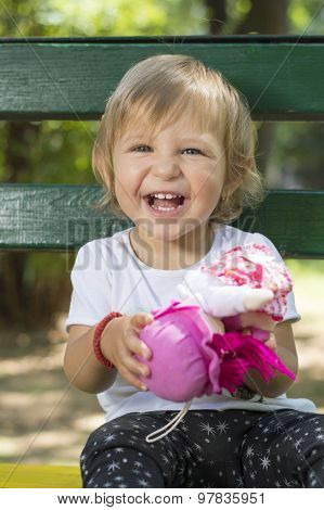 Adorable One Year Old Baby Girl Sitting On A Bench With A Doll In Her Hands