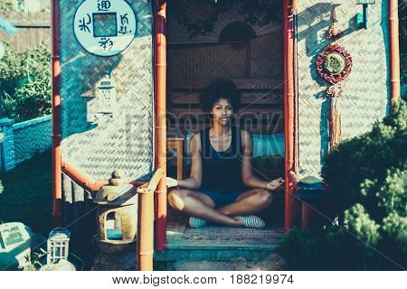 Young charming black girl is sitting cross-legged in Chinese pagoda and practicing relaxing yoga exercises cute curly afro american teenage female enjoying her leisure inside of small asian shack