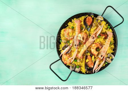 A photo of a Spanish seafood paella in a typical paellera, with mussels, clams, and shrimps, with a place for text, shot from above on a teal texture