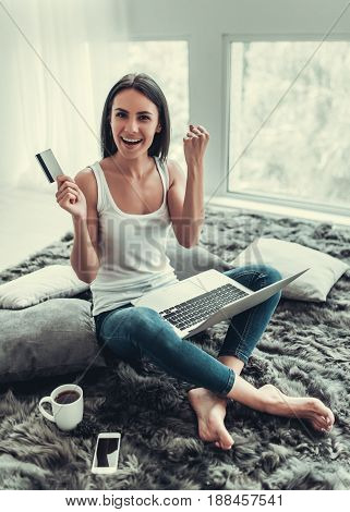 Happy young woman is working with a laptop at home holding a credit card and showing fist