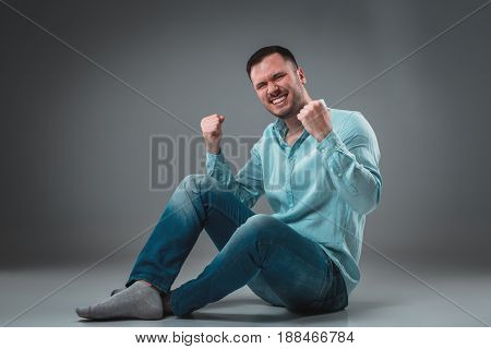 Handsome young man sitting on a floor with raised hands gesturing happiness on gray background. A man in jeans and a blue shirt