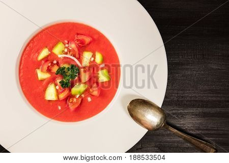 A closeup of a plate of gazpacho soup with slices of tomato, cucumber, and onion, with parsley leaves, on a dark background with a place for text