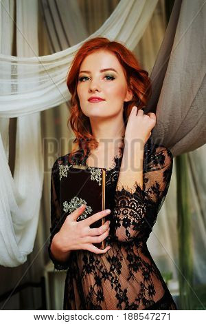 Redhead woman in black lace peignoir on the bed in her bedroom among velvet pillows holding a mysterious black book.