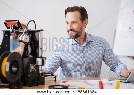 Computer design. Cheerful positive nice man working on the computer and looking at the 3d printer while thinking about a new design