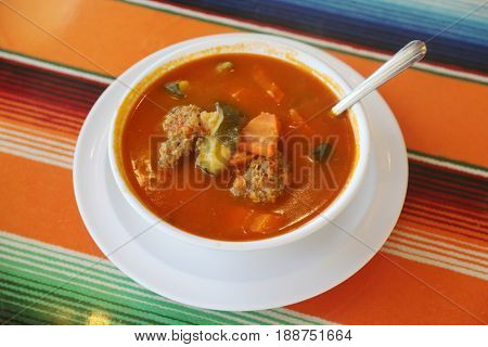 Traditional Mexican food mexican soup with meat balls and vegetables. Mexican  albondigas soup in a white ceramic bowl and plate on a traditional Mexican serape blanked table cloth and a spoon. 