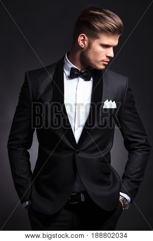 elegant young fashion man in tuxedo looking to his side while holding his hands in his pockets .on black background
