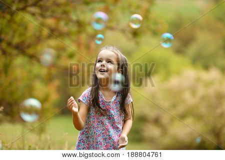 Cute little girl on a forest glade among lush vegetation and around fly soap bubbles. Kid wih soap bubbles over nature outdoor.