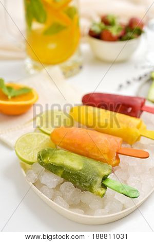 closeup of some different homemade natural ice pops served in a plate with ice, placed on a table with a jar of juice and a bowl with strawberries