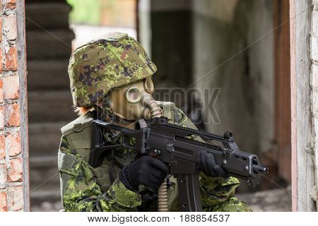 Portrait of armed woman with camouflage. Young female soldier observe with firearm. Child soldier with gun and gas mask in war hearth house ruins background. Military army people concept