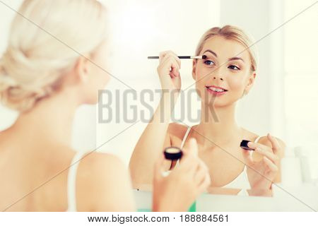 beauty, make up, cosmetics, morning and people concept - young woman applying eyeshade with makeup brush and looking to mirror at home bathroom