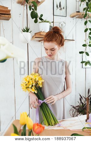 Redhead young florist bandaging yellow bouquet of flowers with violet ribber in workshop