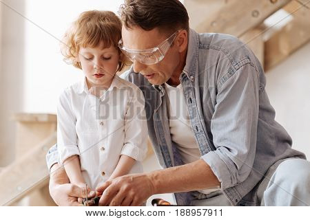 Working mood. Serious father wearing protective glasses and holding pliers in both hand while embracing his son