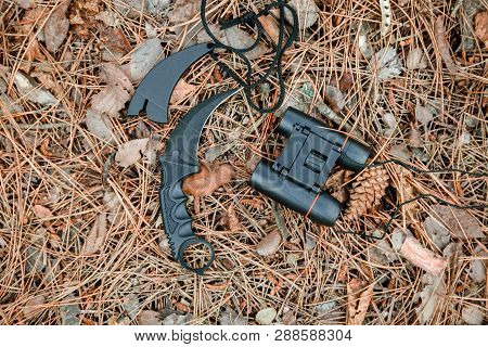 Binoculars And Karambit Knife On The Ground Covered With Pine Needles