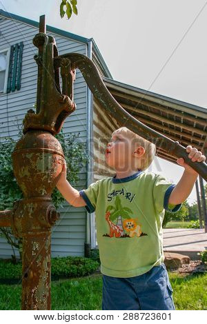 Young Boy Exploring How A Rusted Antique Water Pump Works By An Old Log Cabin