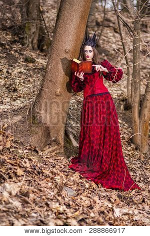 Art Photography. Gorgeous Mysterious Fairy Princess In Red Dress And Black Crown With Old Book. Posi