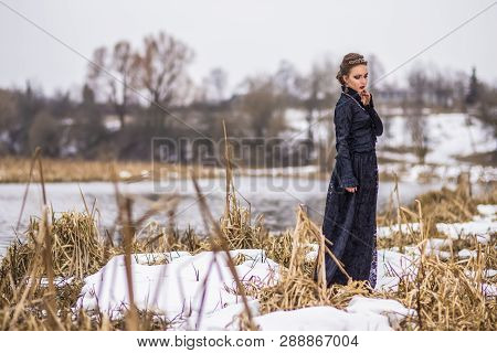 Young Caucasian Brunette Female In Art Dress With Necklace And Crown Posing Against River Outdoors. 